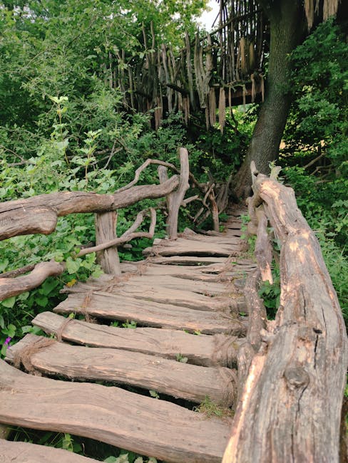 A rustic wooden footpath meanders through a lush, green garden area, featuring uneven, weathered wooden planks forming a narrow walkway. The path is bordered by a handcrafted, uneven wooden railing on