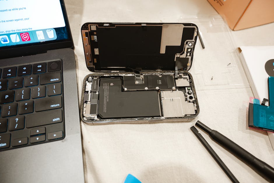 A close-up view of a disassembled smartphone placed on a work surface, with the top screen partly detached and displaying internal components such as the battery and circuit boards. Next to the phone 